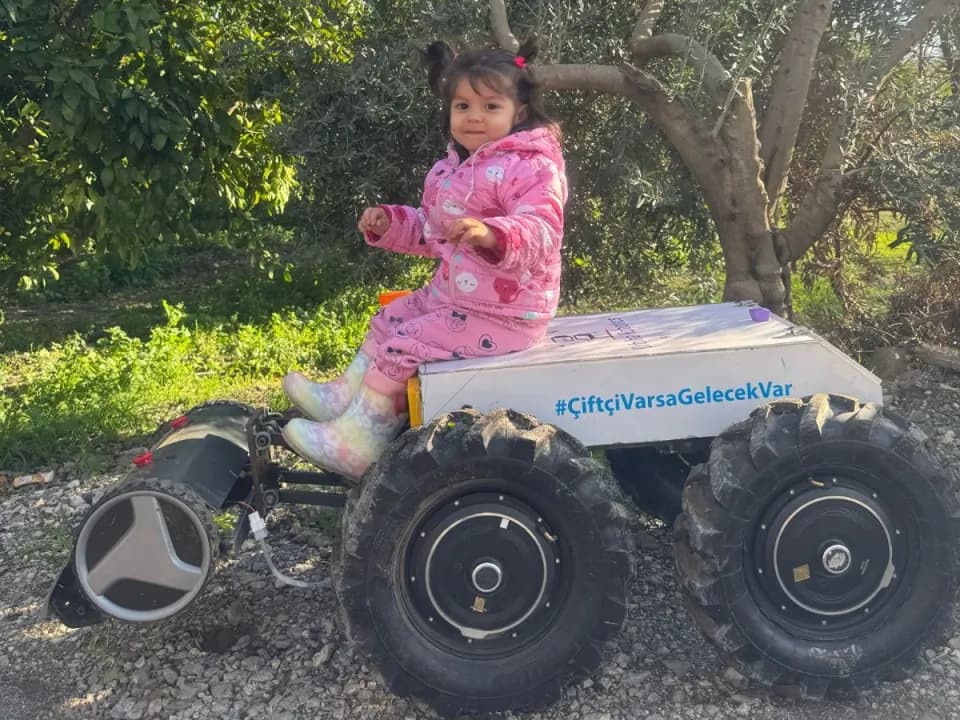 A young child sitting on an Orbiba agricultural robot beneath olive trees